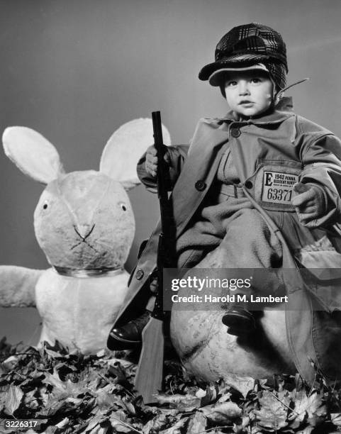 Little boy in a hunting outfit and holding a toy rifle poses on a pumpkin next to a large stuffed rabbit.