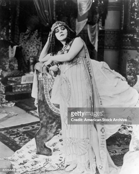 American actor Theda Bara, wearing a costume with entwined sequined snakes and a headdress, leans on a sofa in a still from director J Gordon...