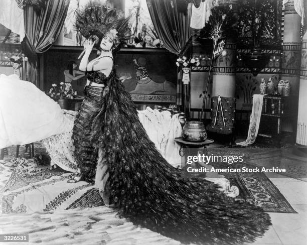 American actor Theda Bara holds a peacock feather fan next to a bed in a still from director J Gordon Edwards' film 'Cleopatra'. Bara is wearing a...