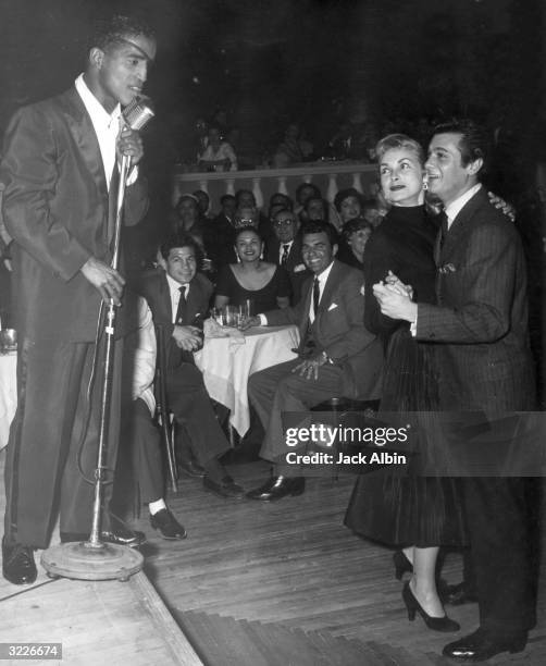 American singer and actor Sammy Davis, Jr. Sings on stage at a microphone, as married American actors Janet Leigh and Tony Curtis dance, at Ciro's...