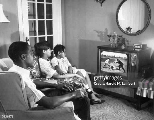 An African-American family sits in their living room, watching a baseball game on television. The mother holds their daughter on her lap.