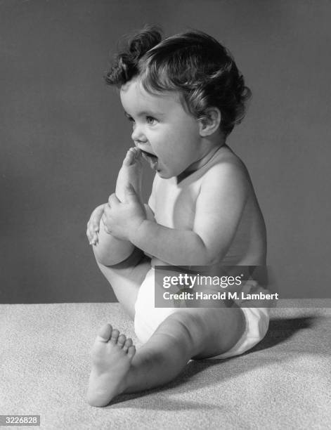 Full-length studio image of an infant sitting on a rug and putting her foot by her mouth. She wears a diaper.