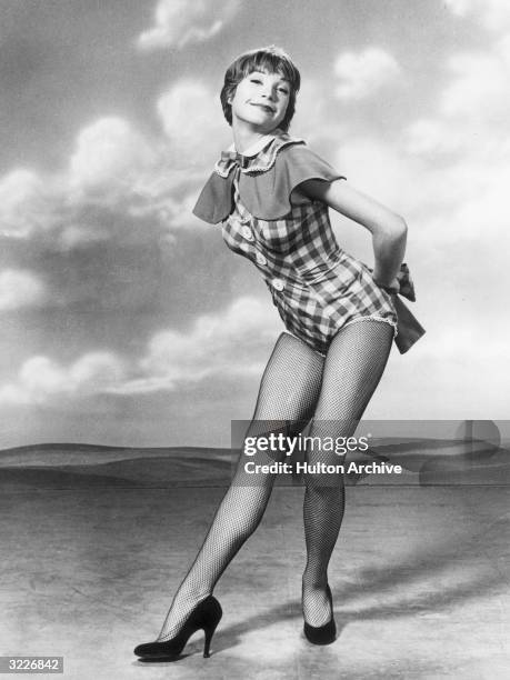 Full-length studio portrait of American actor and dancer Shirley MacLaine, posing in front of a sky backdrop, wearing fishnet stockings and a...