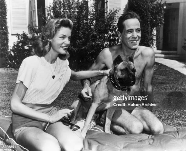 American actor Humphrey Bogart smiles as he kneels with his wife, actor Lauren Bacall, and their pet dog, on a cushion on their front lawn. Bogart...