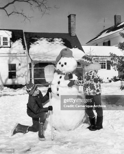 Boy and a girl building a snowman in their front yard. The snowman has a stocking cap, a pipe, a scarf and lumps of coal for its face and buttons.