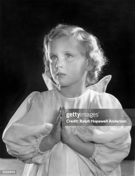 Studio portrait of a young girl holding her hands in prayer with angel wings on the back of her gown, in front of a dark background.