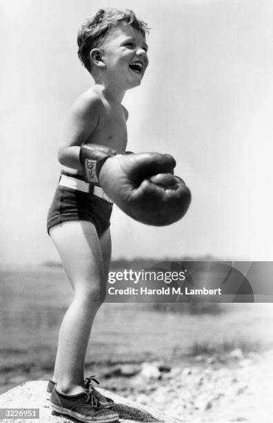 Full-length image of a young boy standing and laughing on a rock by the seashore, wearing adult boxing gloves and shorts.