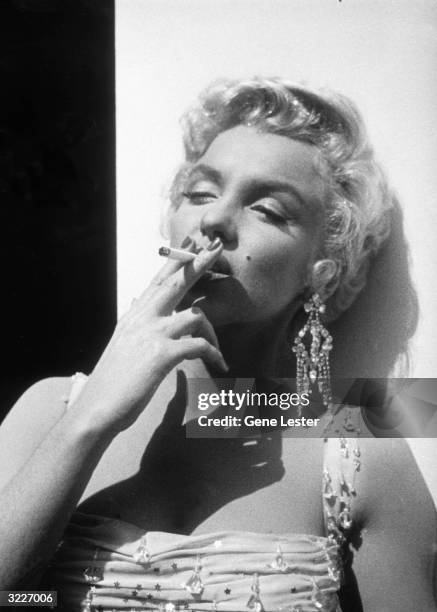 Portrait of American actor Marilyn Monroe in a white beaded gown leaning against a wall smoking a cigarette, while on the 20th Century Fox studios...