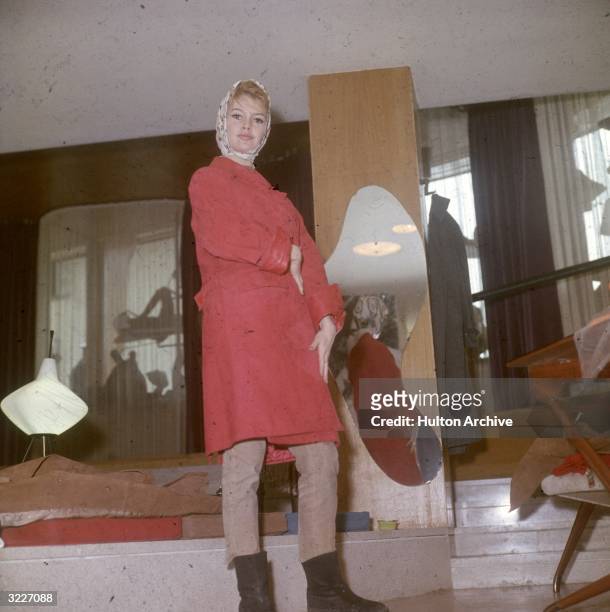 Full-length image of French actor Brigitte Bardot wearing a red suede coat, beige cords, and a silk scarf over her head.
