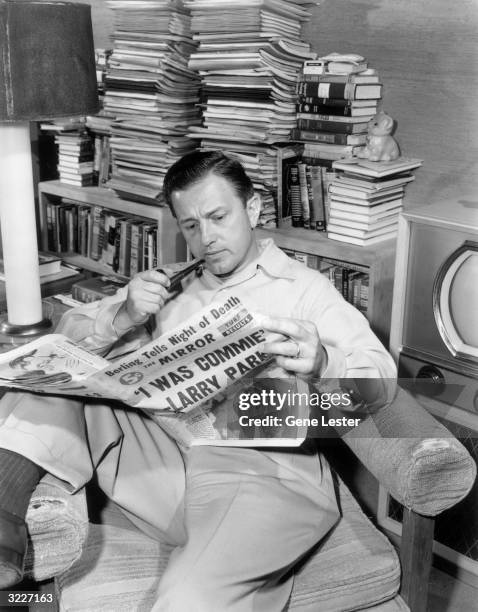 Portrait of American director Edward Dmytryk smoking a pipe while reading 'The Mirror' in a living room, 1950s. The newspaper's headline reads, ''I...