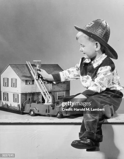 Boy wearing a straw fireman's hat plays with a toy fire engine and a doll house.