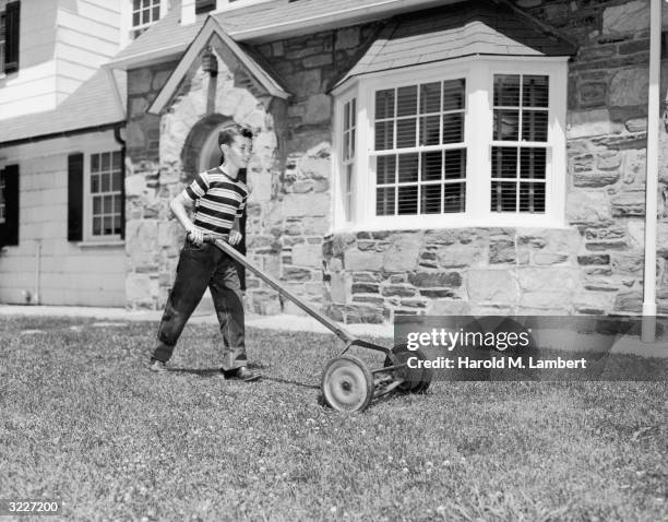 Full-length image of a boy mowing the front lawn of a stone house using a push mower. He wears a striped shirt.