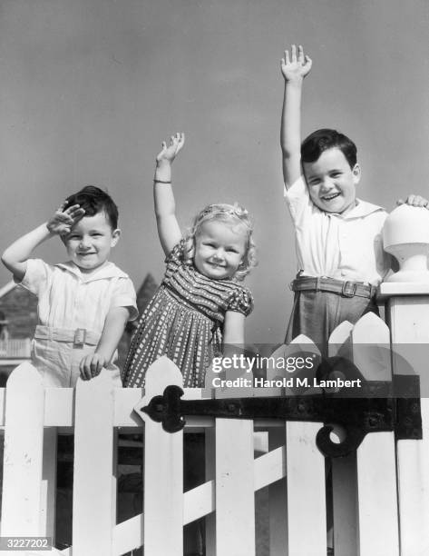 Two young boys and a girl stand behind the gate of a white picket fence, smiling, waving and saluting to the camera.