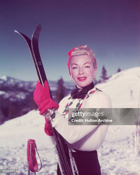 Portrait of American actor Lana Turner , wearing a skiing outfit consisting of a turban, gloves, a knit sweater and ski pants, holding skis on a...