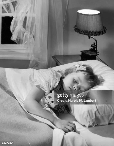 Young girl lies asleep in bed, with a Raggedy Ann doll under her arm. The light on her bedside table is lit.