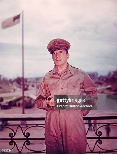 American general Douglas MacArthur , in uniform, holds a pipe while standing on balcony with an American flag in background during World War II.