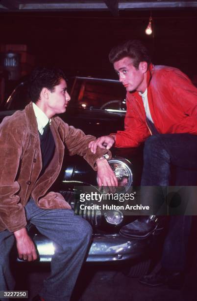 American actor James Dean stands with his foot on the front fender of a car while touching actor Sal Mineo's arm in a still from director Nicholas...