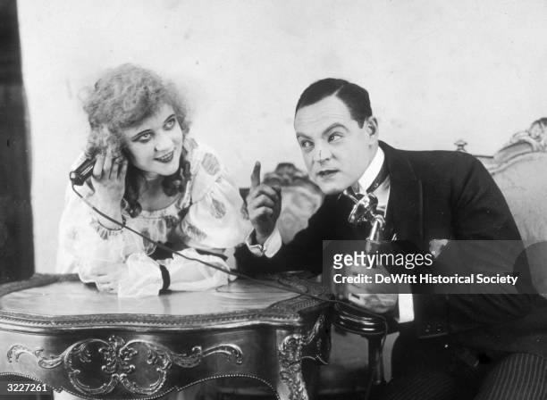 American actors Jean Southern smiling and Howard Estabrook pointing, each holding different parts of a telephone while sitting at a table in a still...