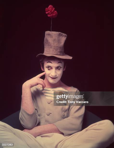 Studio portrait of French actor and pantomimist Marcel Marceau, in costume, sitting cross legged with his finger on his cheek and an artificial...
