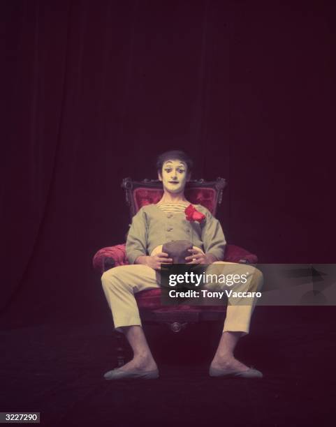 Studio portrait of French actor and pantomimist Marcel Marceau, in costume, sitting in a wooden arm chair with his hat and an artificial flower in...