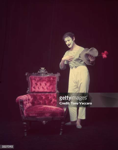 Full-length studio portrait of French actor and pantomimist Marcel Marceau, in costume, standing beside an arm chair with a flower in hand and...