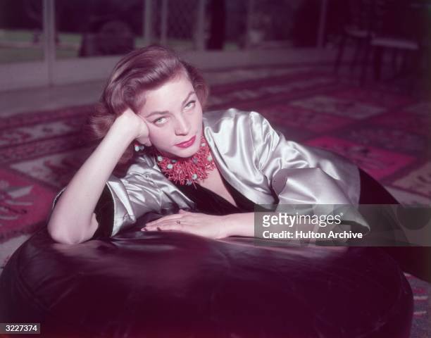 Portrait of American actor Lauren Bacall wearing a coral necklace and earrings and a gold satin jacket over a black velvet body suit, seated on an...