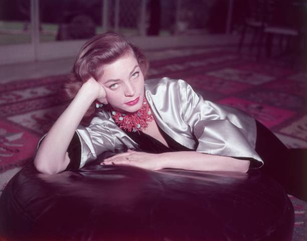 Portrait of American actor Lauren Bacall wearing a coral necklace and earrings and a gold satin jacket over a black velvet body suit, seated on an...