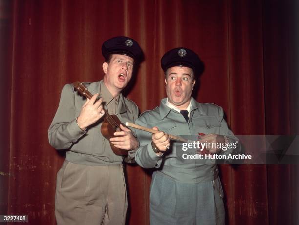 Portrait of American comedy team, actors and entertainers Abbott and Costello, dressed in blue collar uniforms, Bud Abbott playing a ukulele and Lou...