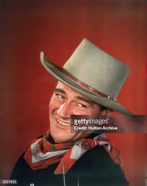 Studio headshot portrait of American actor John Wayne smiling in front of a red background, dressed in Western garb, with his head turned to the side.