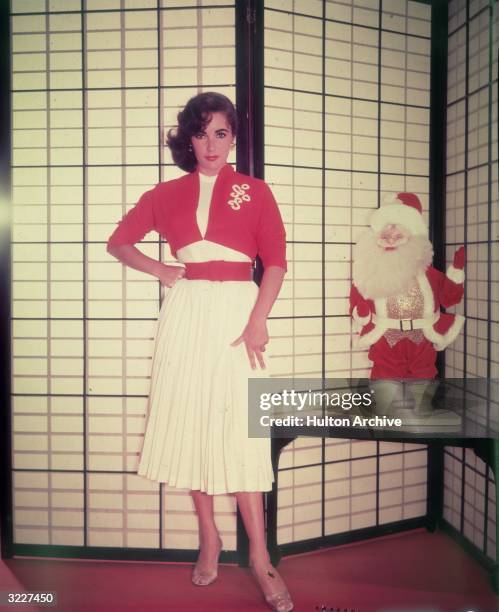 Promotional portrait of British-born actor Elizabeth Taylor standing next to a Santa Claus doll in front of a shoji screen. Taylor wears a red bolero...