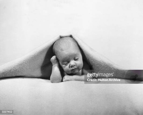 Newborn baby with its head protruding from under a blanket, leaning against its hand.