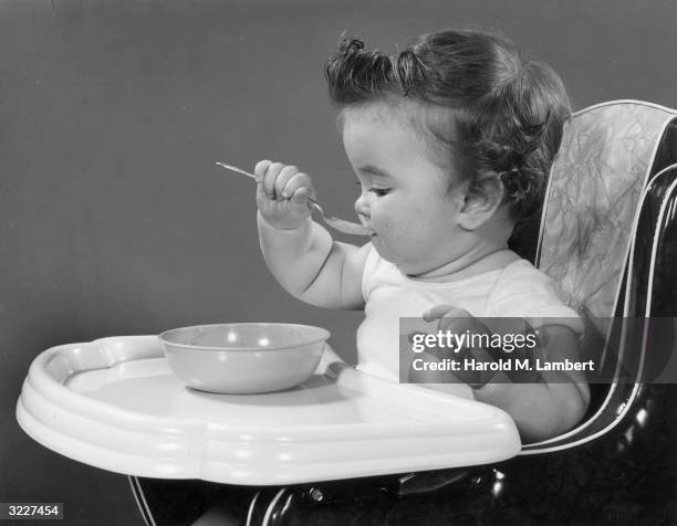 Baby sits in a high chair, eating from a bowl with a spoon to her mouth.