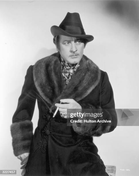 American actor John Barrymore holds a cigarette while wearing a costume consisting of a hat, an ascot, and a dark coat with a fur collar and cuffs in...