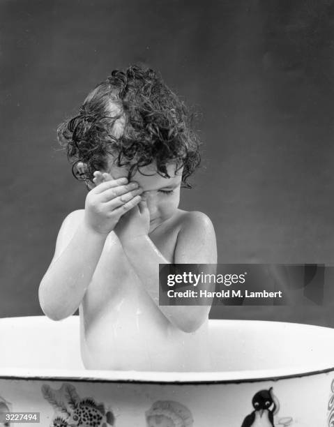 Studio image of a female infant sitting up in a bathtub, and holding her hands over one eye.