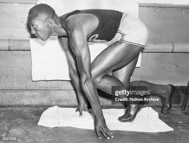 American track and field athlete Jesse Owens crouches in starting position while preparing for a race. He is wearing a singlet, shorts, and cleated...