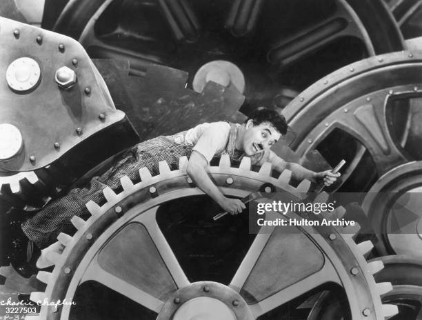 British actor and director Charles Chaplin , wearing overalls and holding a wrench, sits on an enormous set of gears in a still from Chaplin's film,...