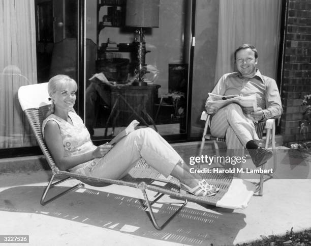 Portrait of a couple smiling as they sit in lawn chairs on their backyard patio. The man sits with a newspaper, while the woman reclines with a book.