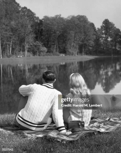 Man and woman sit on a blanket in the grass, with their backs to the camera, looking out over a lake. They have a small radio on the blanket.