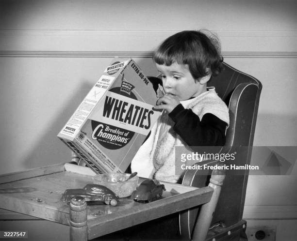 Young girl sits at a table, digging with one hand in a box of Wheaties breakfast cereal. There is an empty bowl and two toy cars on the table.