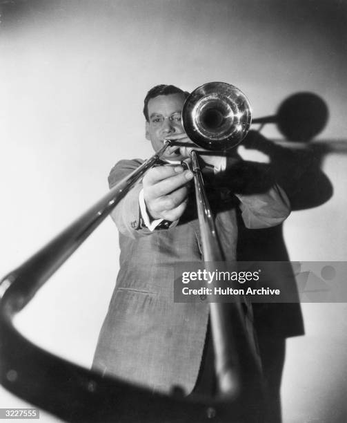Low-angle studio portrait of American bandleader and musician Glenn Miller playing a trombone with the slide in the immediate foreground.