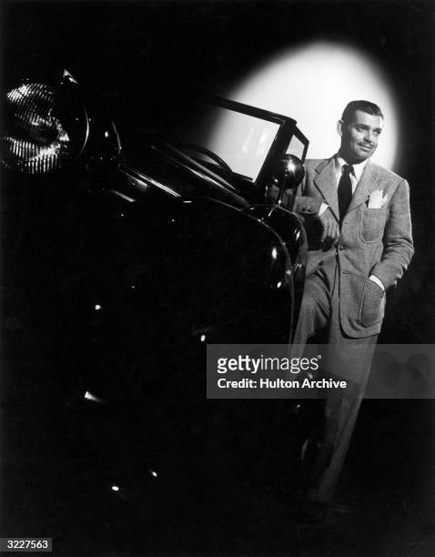 Studio portrait of American actor Clark Gable leaning against a car in a spotlight. Gable is wearing a suit and tie.