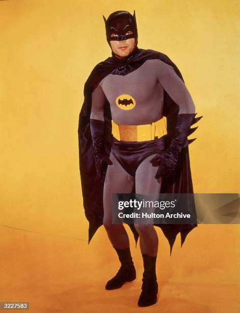 American actor Adam West poses in costume as Batman in front of a yellow backdrop in a promotional portrait for the television series, 'Batman'.