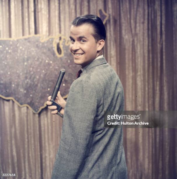 American TV host and producer Dick Clark looks over his shoulder while holding a microphone on the set of the television series, 'American Bandstand'.