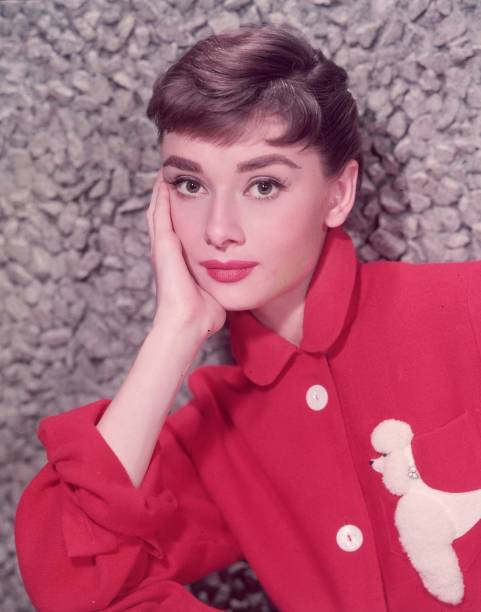 Headshot portrait of Belgian-born actor Audrey Hepburn leaning on her hand in a red jacket with a poodle applique.