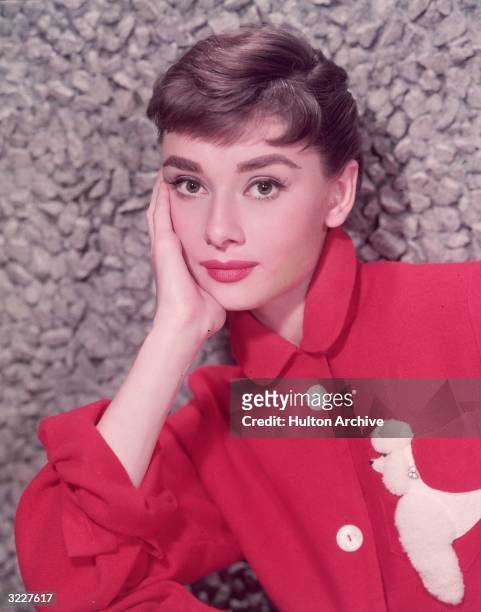 Headshot portrait of Belgian-born actor Audrey Hepburn leaning on her hand in a red jacket with a poodle applique.