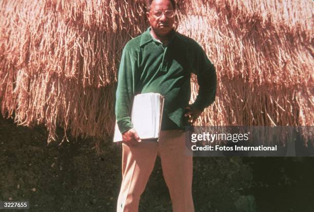 American writer Alex Haley holds a script as he stands in front of a grass hut on the set of the television mini-series, 'Roots,' based on his novel...