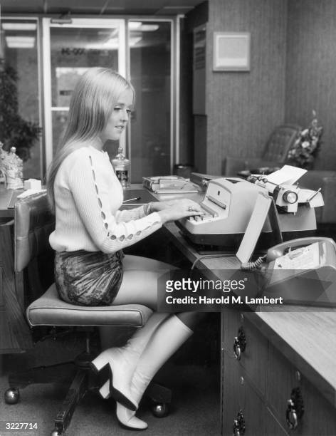 Teenage girl smiles while sitting at an office desk, typing on an electric typewriter. She wears a miniskirt and knee-high boots.