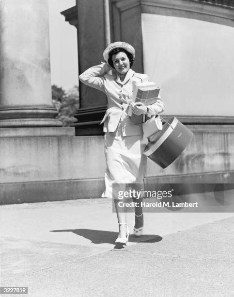 Woman smiles as she walks down a sidewalk, carrying several wrapped packages and a hatbox. She has one hand on top of her hat.