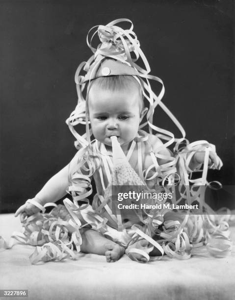 Studio image of a baby sitting and blowing a noisemaker, with streamers draped over him. He wears a party hat.