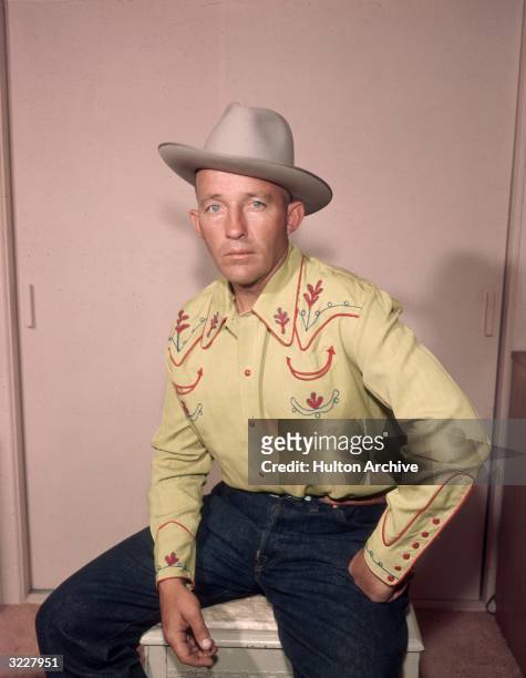 Studio portrait of American actor and singer Bing Crosby seated with his legs spread apart and his fist on his hip, wearing a western shirt, jeans,...